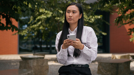 Fototapeta premium Asian Korean business woman holding mobile phone smartphone thinking message chatting online social media businesswoman girl Chinese Japanese thoughtful female at city outdoors lady find answer idea