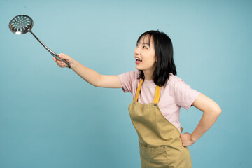 Cheerful Asian woman in an apron happily holds a cooking utensil while posing against a vibrant...