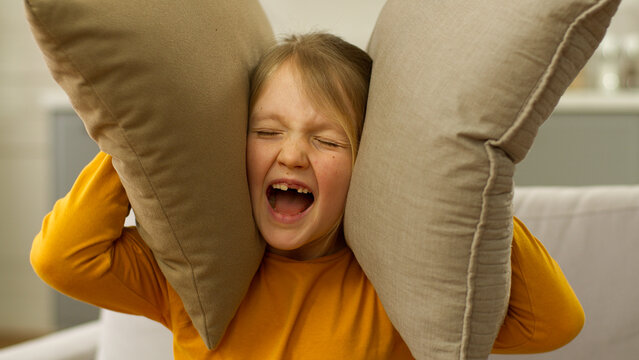 Little Caucasian girl child screaming yelling shouting loud covering ears with pillows protest education daughter offspring displeased anger emotional furious distraught aggression dramatic preteen
