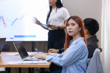Women with red hair attentively works on her laptop while colleague delivers presentation.