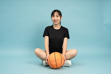 A young Asian woman sits on the floor with one leg crossed and a basketball beside her, smiling and...
