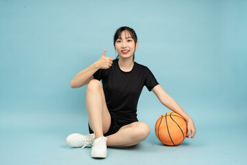 A young Asian woman sits on the floor with one leg crossed and a basketball beside her, smiling and...
