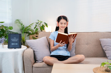Smiling young asian woman siting in room enjoying her favorite music in headphones, reading book, ...