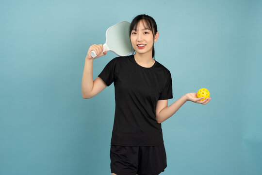 Smiling young Asian woman holding a pickleball paddle and ball against a light blue background, promoting sports and active lifestyle in a playful manner