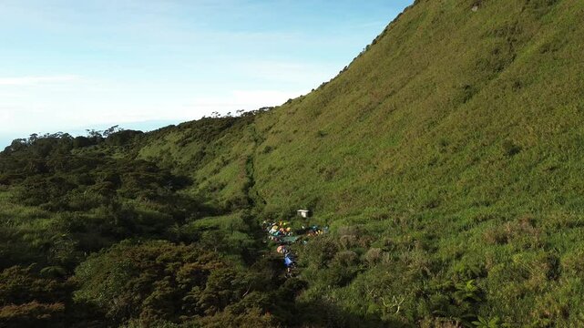 Emerald slopes of Mount Apo arc above a narrow forested ravine where colourful tents and a small hut cluster beside a trickling stream, all bathed in clear dawn light.