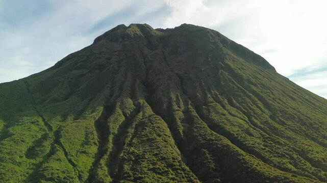 The camera advances toward Mount Apo, exposing radial green gullies carved down its massive cone while soft morning light and wispy summit steam rise into a pale blue sky.