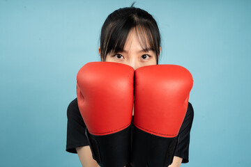 Young Asian person in black shirt holds red boxing gloves close to face against a blue background,...