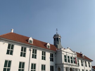 Grand historic white building with a red tile roof, classic architectural details, dormer windows, and a central cupola against a clear blue sky, s...
