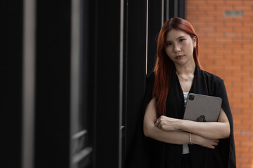 Confident woman with long red hair leans against modern wall, holding tablet closely to her chest....