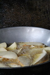 Slices of cassava are deep fried in hot oil inside a traditional wok over a gas stove. A simple street food scene in a rustic kitchen with natural lighting and rough wall background.
