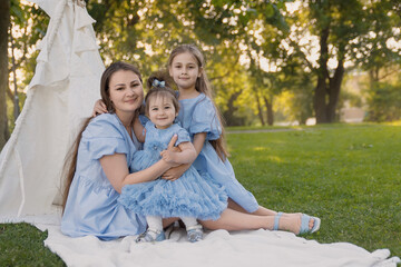 Seven, a mother and her daughters are outdoors, sitting on a blanket in the park. All of them are...