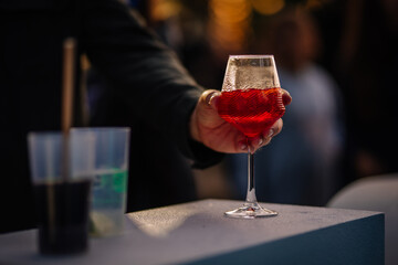 Close-up of a hand holding a red cocktail in a textured wine glass at a silent disco, with plastic cups and a blurred crowd in the background.