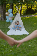 Two sisters are dressed in a blue dress, playing in a teepee in the park, with balloons in the...