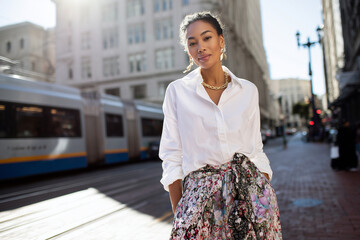 Woman wearing classic white long sleeve shirt tucked into flowy printed midi skirt, standing in sunny urban street, 