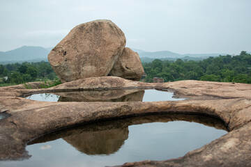 A massive boulder perches on a rocky hilltop, its reflection captured in rain-filled pools. Lush...