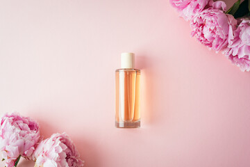 Glass cosmetic perfume bottle surrounded by pink peony flowers on pastel pink background. Top view, flat lay