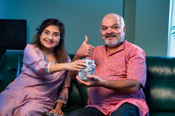 Indian senior couple promoting saving money by putting note in glass jar sitting at home