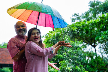 Elderly Indian couple sharing joyful rainy moment outdoors under umbrella during monsoon