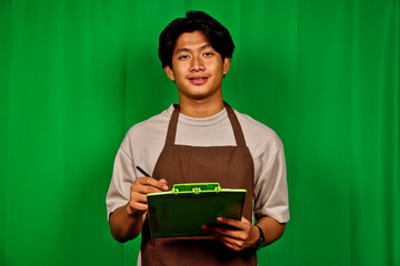 Young Man Wearing Apron Standing in Front of Green Screen Holding Clipboard and Pen