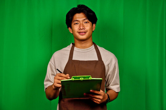 Young Man Wearing Apron Standing in Front of Green Screen Holding Clipboard and Pen