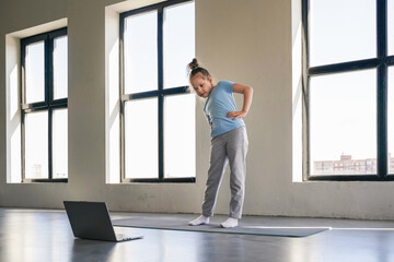 A young girl performs yoga stretches in a well-lit room while following instructions from a laptop