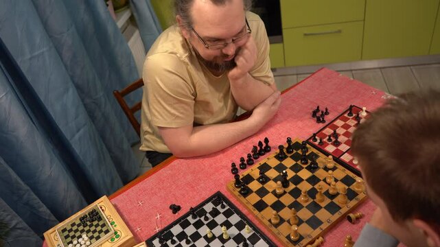 Father and son play chess on three boards at once. Teenage boy plays with dad. Family time together at home. - Powered by Adobe