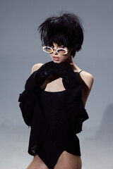 young woman wearing black bodysuit and gloves with white sunglasses posing against gray background,...