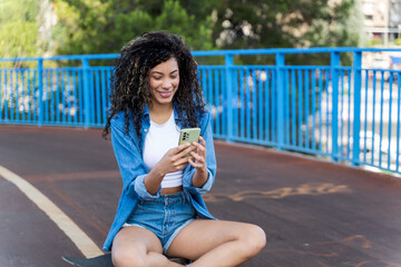 Smiling young woman using mobile phone while sitting on skateboard on bridge in the city