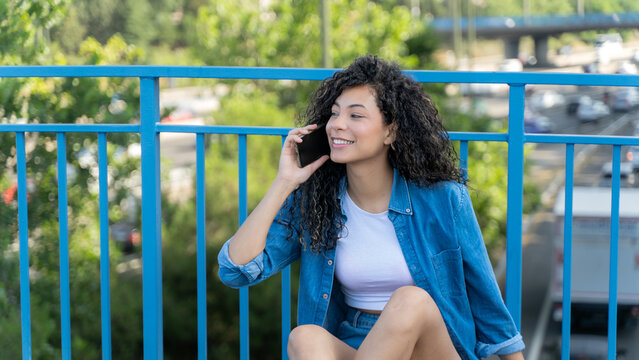 Smiling young woman talking on smartphone while sitting on skateboard on bridge in city - Powered by Adobe