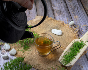 Close-up of a man’s hand holding a glass cup of horsetail tea over rustic wood and herbs. Natural...