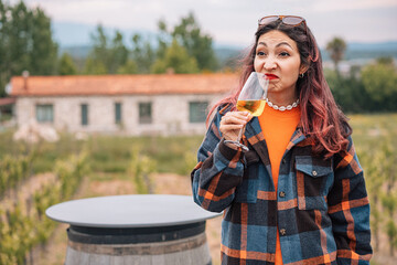 Woman grimacing while drinking a glass of bad white wine in a vineyard with a farmhouse in the...