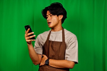 Young man in casual beige and brown apron talking on a mobile phone with a thoughtful expression in front of a vibrant green background, ideal for communication or food service marketing.
