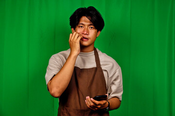 Young Man Wearing Brown Apron and Casual Clothes Standing in Front of Green Screen Holding Smartphone