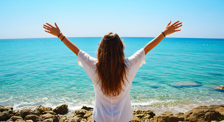 A woman with long hair in a white shirt stands with arms raised on a rocky beach near turquoise ocean under a clear blue sky