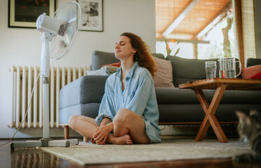 Young woman sitting at home on the floor in front of a fan saving from heat