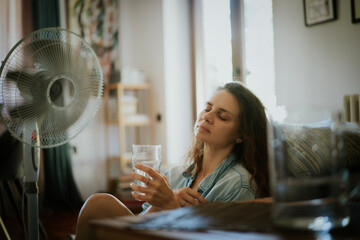 Young woman sitting at home on the floor in front of a ventilator with glass of water , saving from...