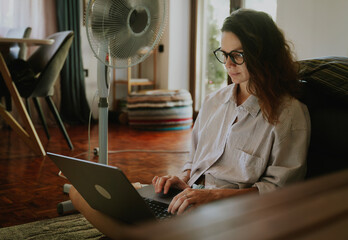 Beautiful young woman with laptop and electric fan sitting on the floor in living room