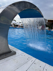 Modern outdoor swimming pool with waterfall fountain, people relaxing in background, summer leisure concept.