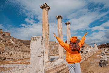 Young woman tourist is posing with raised arms near ancient ruins of Asklepion, famous landmark in...