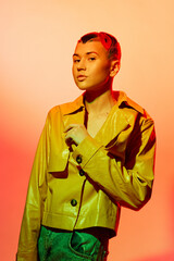 Woman with short hair wearing a glossy yellow jacket and green pants, posing against an orange gradient background, stylish and bold expression.