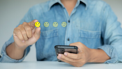 A man using a smartphone to select a sad face icon on a virtual customer feedback rating,...