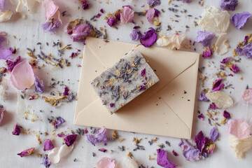 Handmade botanical soap bar with dried flower petals on beige envelope, surrounded by scattered pastel flower petals, creating soft, romantic, and natural flatlay composition