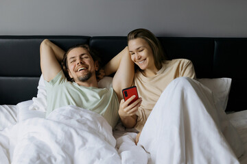 Young caucasian couple relaxing in bed with smartphone, smiling and enjoying leisure time