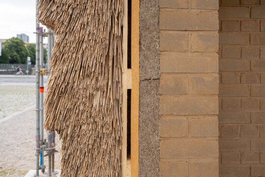 Close-up of a sustainable building wall cross-section showing natural insulation materials including reed, hemp, and clay bricks, at a construction site