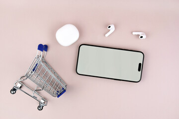 Shopping cart with a smartphone and white wireless headphones on a pink background.