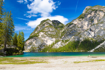 Secluded corner of Lake Braies with its vibrant turquoise water meeting a light-colored shore, framed by towering, rugged mountains and lush green forests under a bright sky.