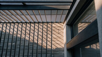 Obraz premium Shadows of a metal railing cast geometric patterns on a textured brick wall and window frame in warm natural light.