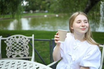 A girl in a white top sits at a table, savoring a warm drink while gazing at a tranquil pond...