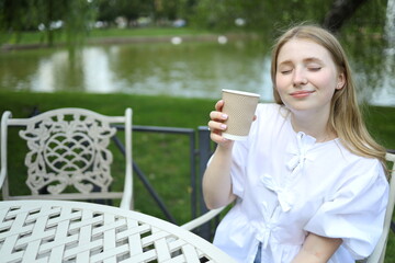 A young woman sits at a white table by a tranquil lake, savoring a warm drink with her eyes closed....