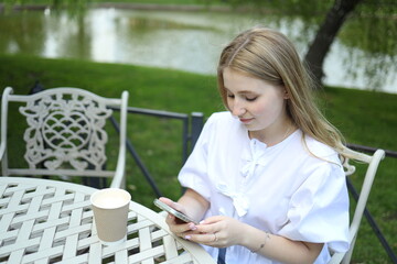 A young woman with long blonde hair relaxes at a garden table, texting on her smartphone while...
